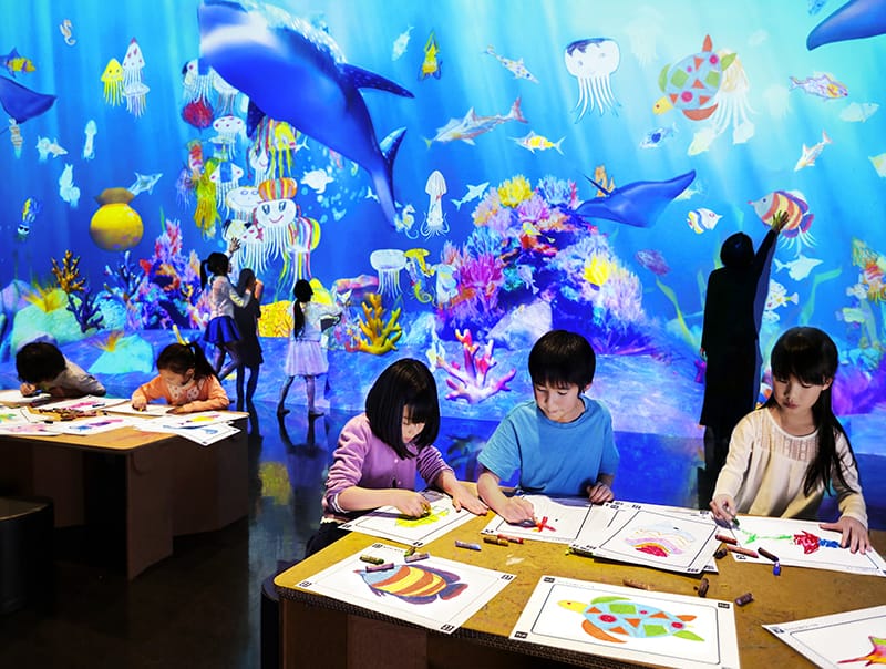 Wide shot of TeamLab installation where kids color ocean creatures that later become animated onscreen